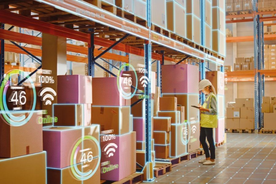 Eine Mitarbeiterin in einem Lager prüft gestapelte Kisten und hält dabei ein Tablet in der Hand. Die Kisten sind von leuchtenden digitalen Overlays umgeben, die Status und Konnektivität anzeigen. Die Mitarbeiterin trägt eine gelbe Weste und einen Helm.